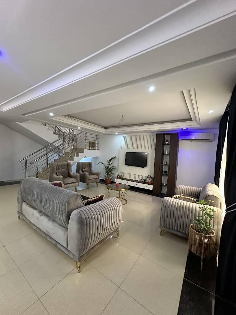 A spacious living area is showcased with modern furnishings, including two plush sofas and four armchairs arranged around a coffee table. A flat-screen television is mounted on the wall, complemented by decorative shelves. Natural light fills the room, highlighting the elegant staircase in the background.