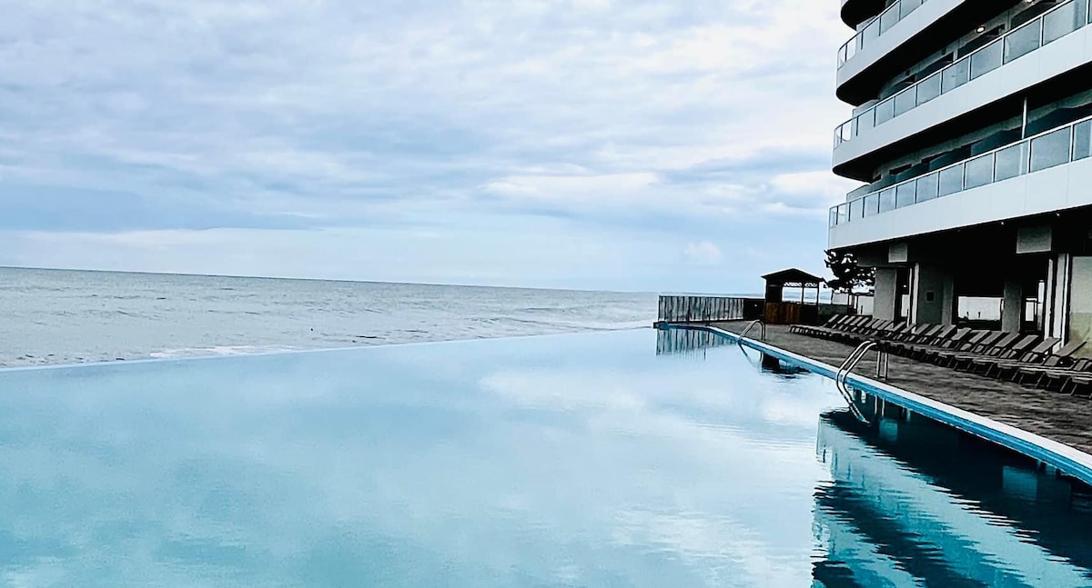 An infinity pool reflects the sky and ocean, creating a tranquil atmosphere. The pool is bordered by sun loungers and overlooks the water. The modern building's sleek lines are visible in the background, enhancing the relaxed coastal setting.