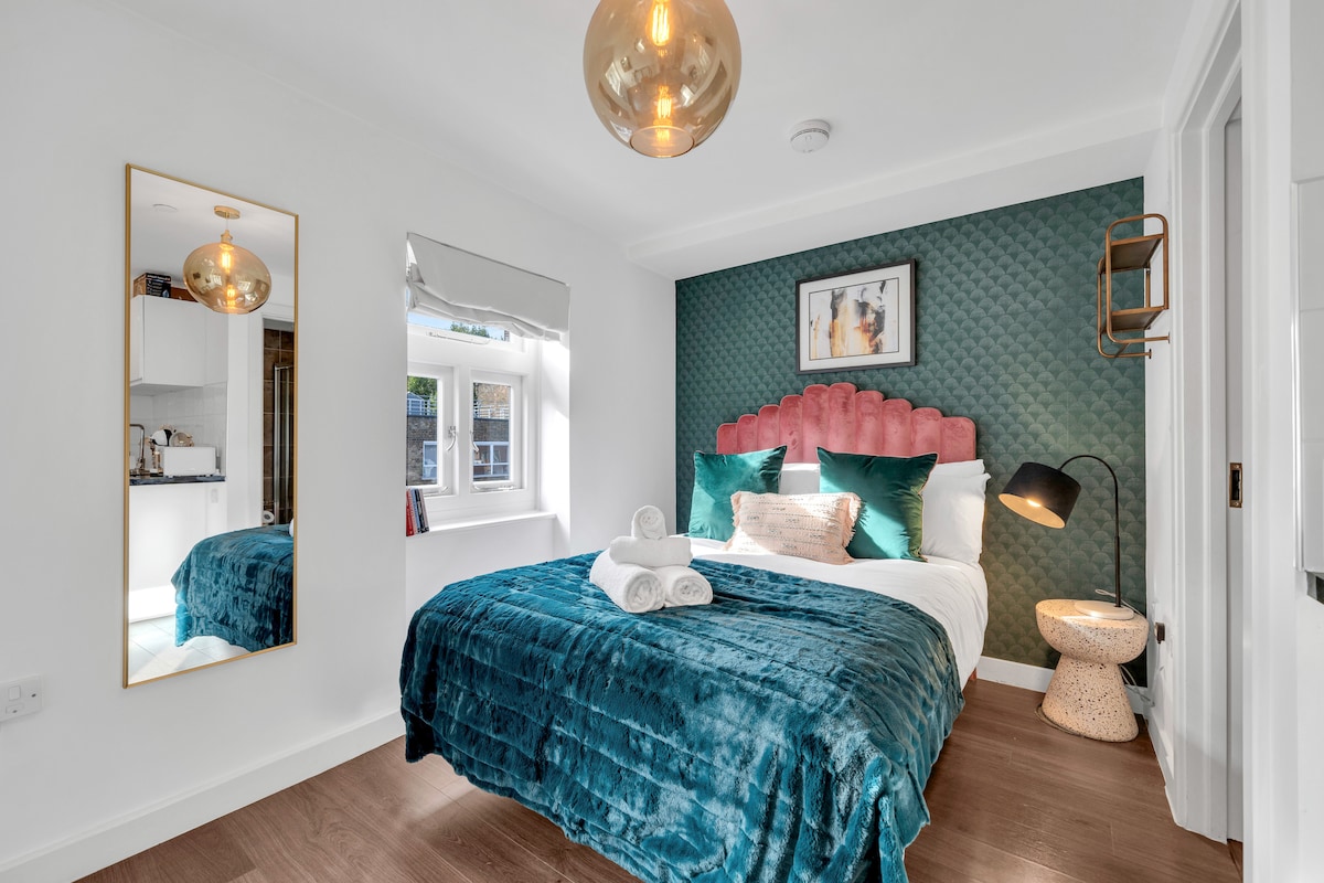 The bright sleeping area features a spacious bed with a pink headboard and decorative pillows. A floor lamp stands beside a small side table, and a mirror reflects the serene space. Natural light fills the room through a window with a blind.