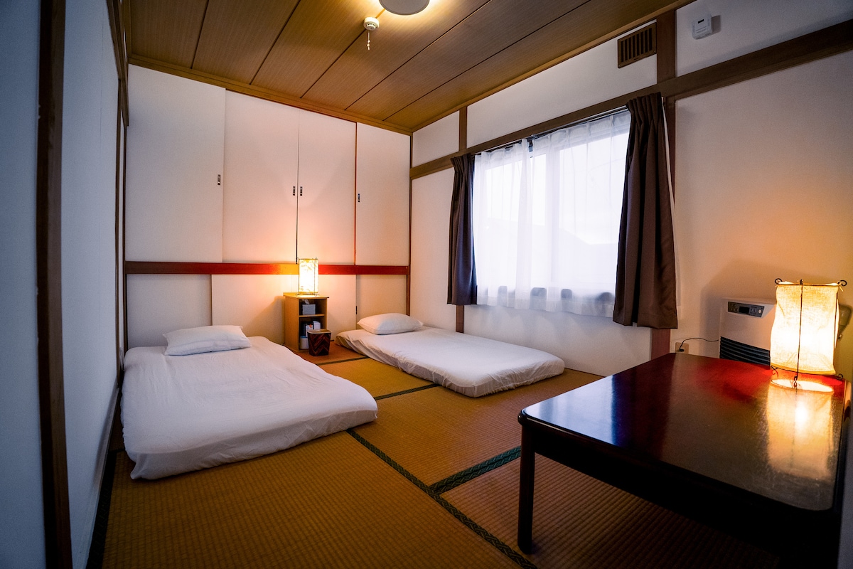 A traditional Japanese room features two futon beds neatly laid on tatami flooring. A small wooden table sits in the center, complemented by two decorative lamps. A window allows natural light to brighten the space, adorned with light curtains for privacy.