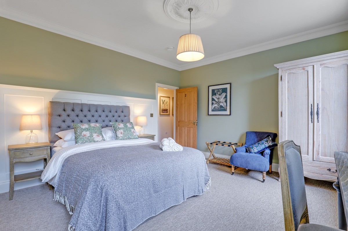 A spacious room is furnished with a large bed dressed in soft linens and decorative pillows. A vintage armoire and a comfortable chair enhance the decor. Soft lighting is provided by wall sconces and a pendant lamp overhead, while a window allows natural light to fill the space.