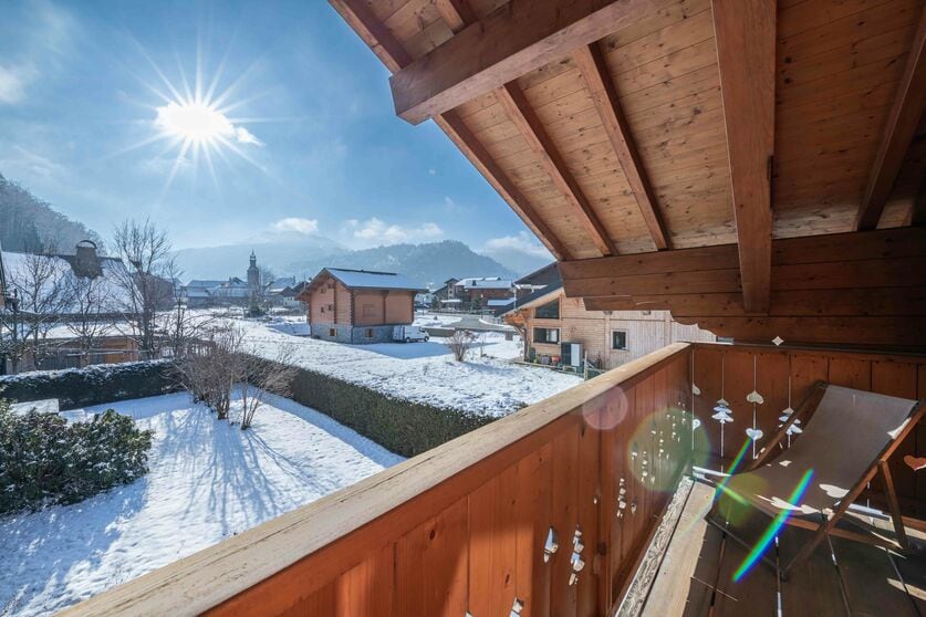 A balcony scene reveals a stunning view of snow-covered landscapes and distant mountains, complemented by sunlight reflecting off the snow. Wooden structures dot the scenery, creating a serene atmosphere. A reclining chair is positioned for relaxation, enhancing the appeal of this outdoor space.
