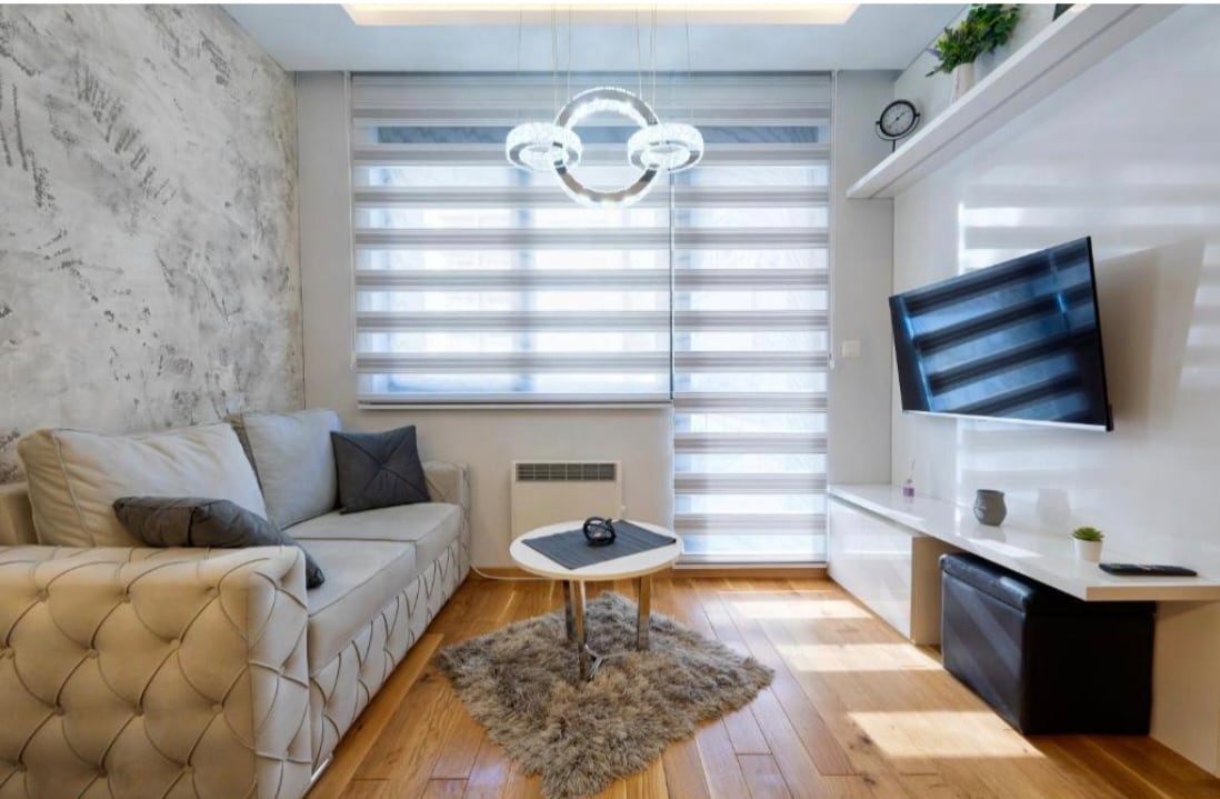 A modern living area features a light-colored, quilted sofa and a small round coffee table on a soft area rug. Large windows are adorned with elegant blinds, allowing natural light to fill the space. A sleek TV unit and decorative plant enhance the contemporary design.