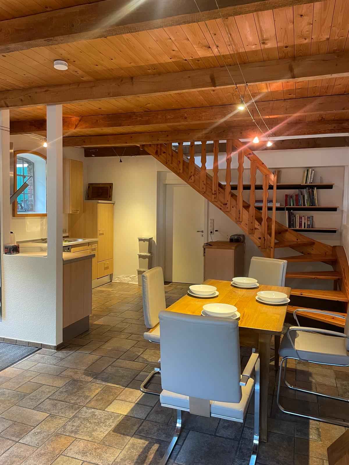 An open-plan living and dining area is shown, featuring a wooden table set for four with white plates. A fully equipped kitchen is visible in the background. Natural light streams in through a window, highlighting the wooden ceiling beams and the staircase leading to the upper floor.