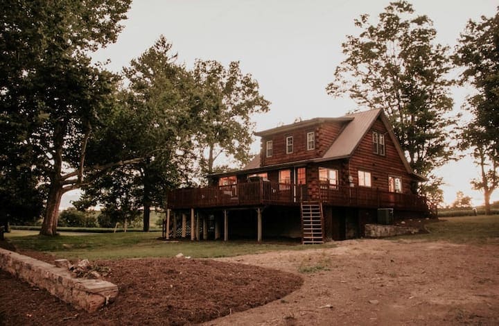 A cozy cabin is surrounded by tall trees, featuring a spacious deck that extends from the front of the home. The two-story structure showcases large windows, allowing natural light to fill the interior. A gravel area is present in the foreground, adding to the outdoor space.