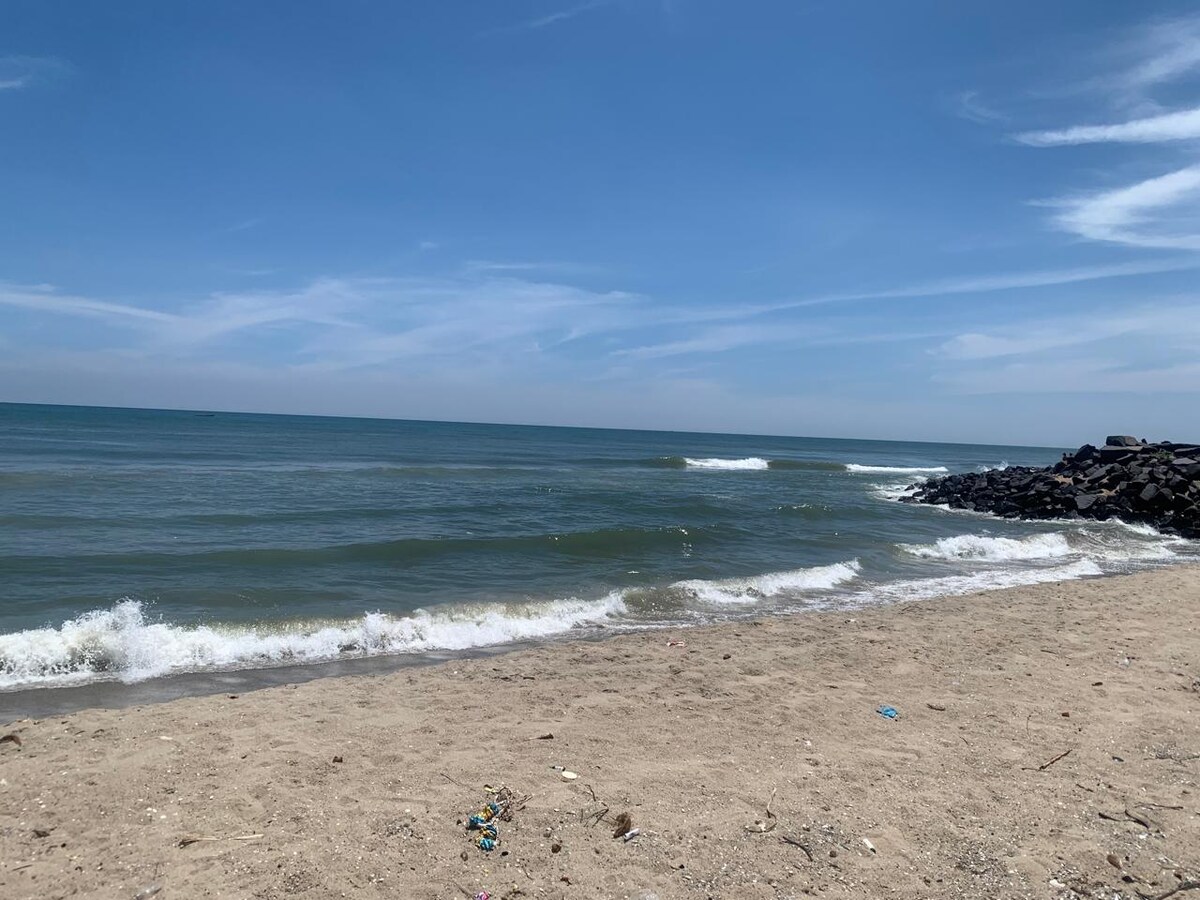 A sandy beach stretches along a calm shoreline, with gentle waves lapping at the water's edge. The ocean showcases varying shades of blue under a clear sky, with wispy clouds scattered above. A rocky jetty is visible on the right side of the image.