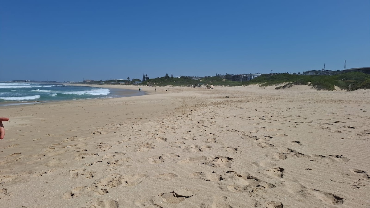A wide sandy beach stretches along the coastline, with gentle waves lapping at the shore. Footprints are visible in the sand, leading towards the water. Low greenery and distant buildings form the background under a clear blue sky.