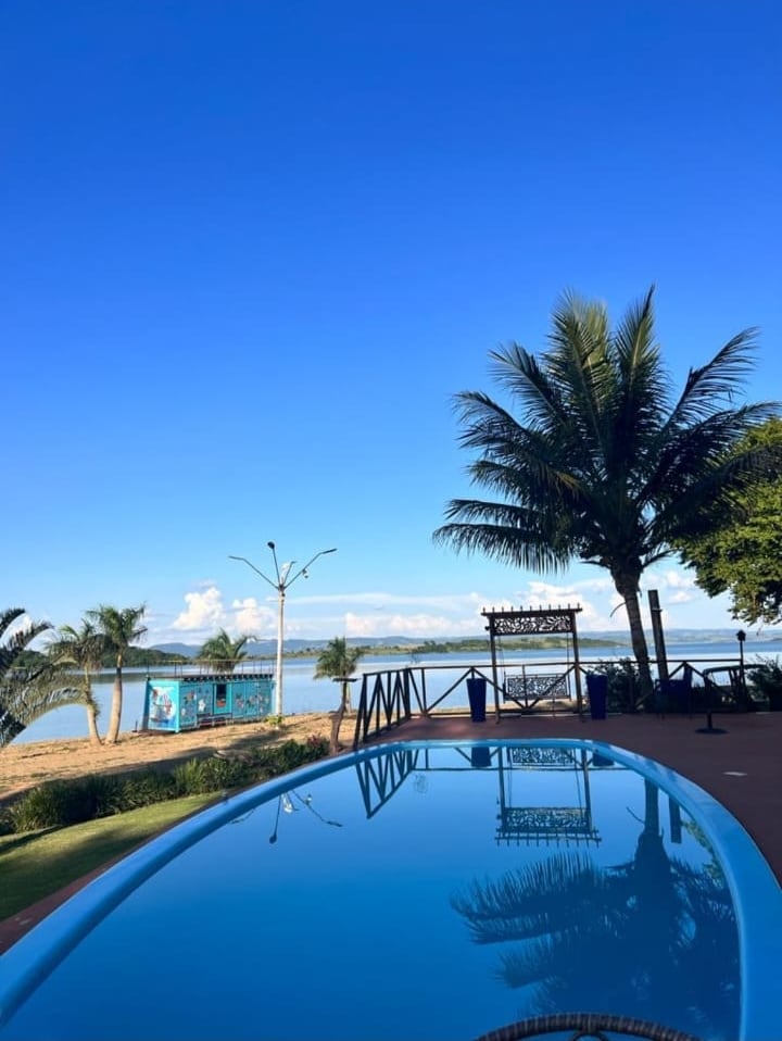 A tranquil pool reflects the clear blue sky, bordered by lush greenery and palm trees. The scenic view of the beach and water is visible in the background, enhancing the serene atmosphere of the outdoor space.