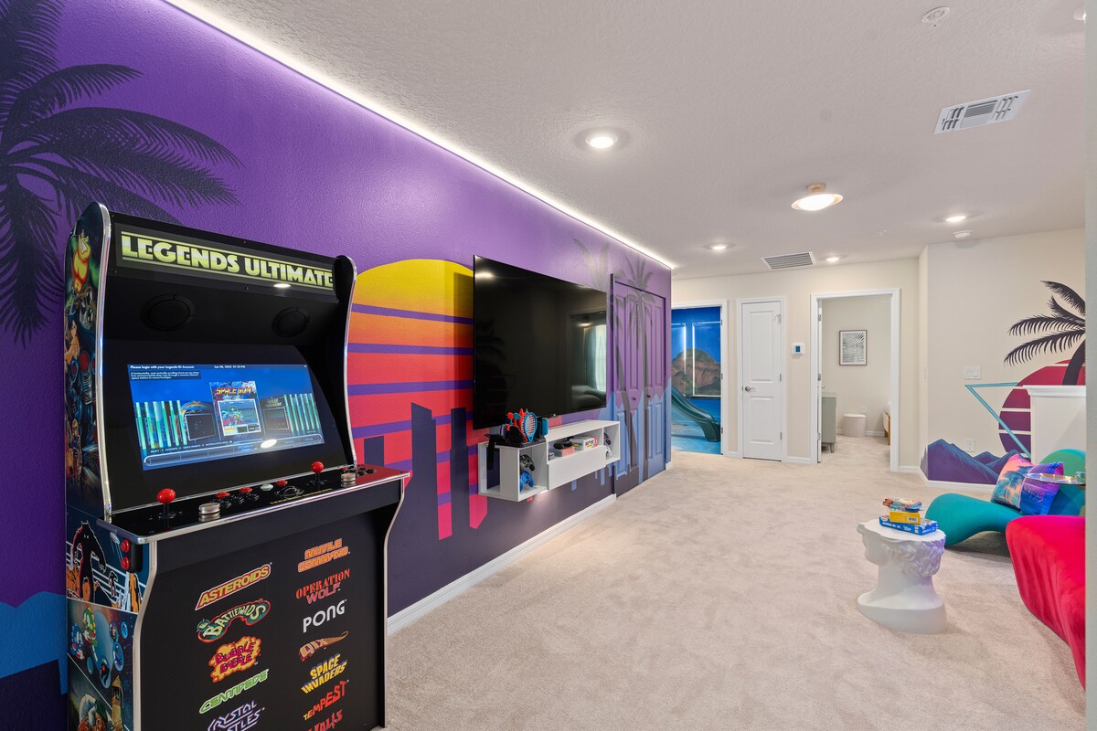 A vibrant game room features colorful murals and soft carpet. An arcade cabinet stands prominently, alongside a large television mounted on the wall. Colorful seating options invite relaxation, and bright lighting enhances the playful atmosphere of the space.