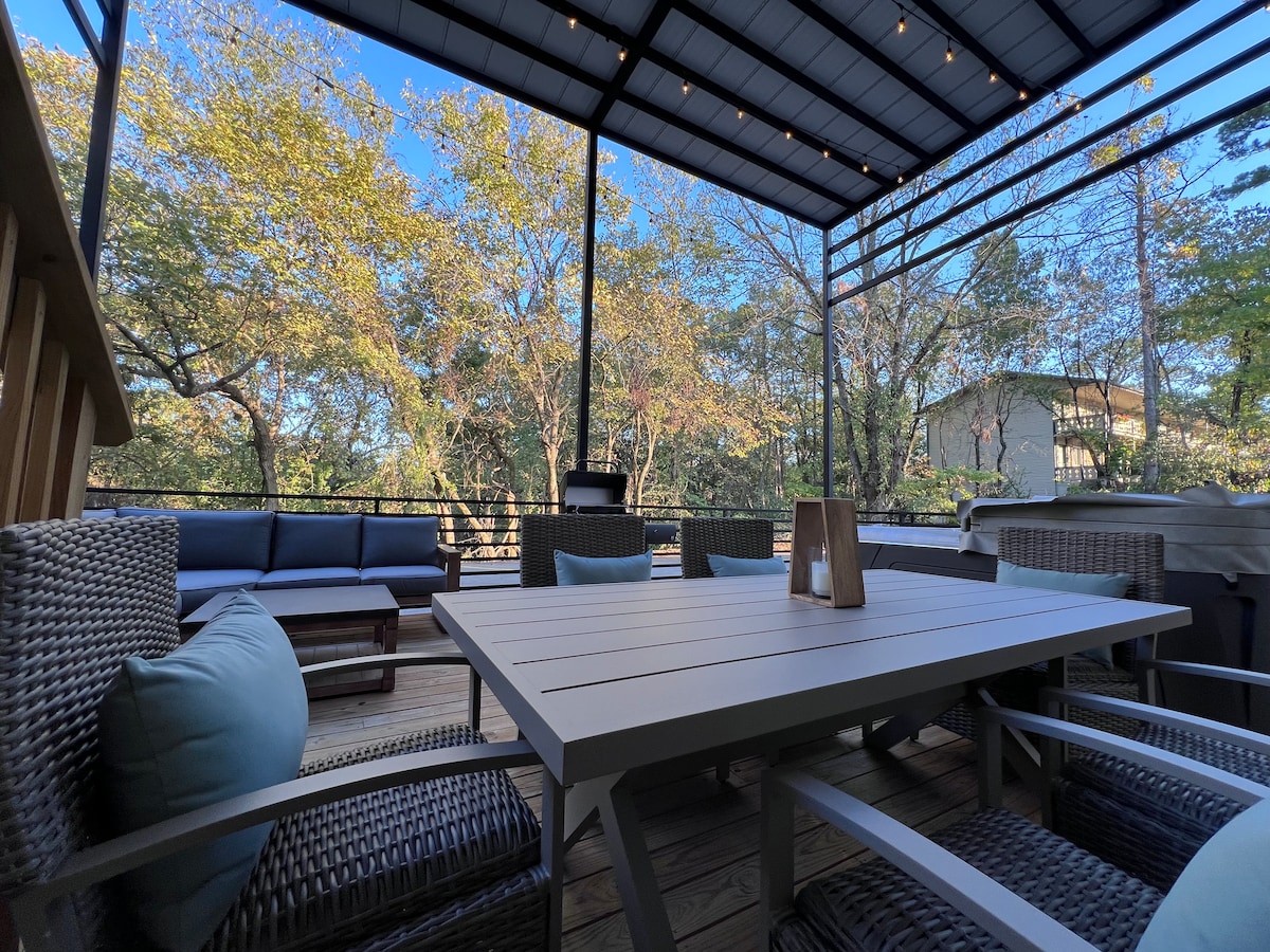 An outdoor patio is presented, featuring a spacious table surrounded by woven chairs with soft cushions. A comfortable seating area with a sectional sofa is visible in the background, and trees provide a natural backdrop under clear blue skies.