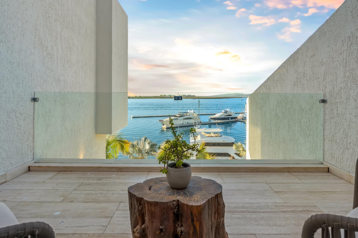 A private terrace offers sweeping views of the marina and the bay, featuring a rustic wooden table topped with a small potted plant. Two comfortable chairs are positioned to enjoy the scenic waterfront and the serene atmosphere of the surroundings.