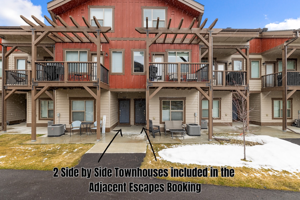 Two side-by-side townhouses are depicted, featuring a modern exterior with wooden accents and balconies. The entrance is flanked by outdoor seating. Snow is visible on the ground, suggesting a colder climate, while the clear blue sky provides a bright backdrop.