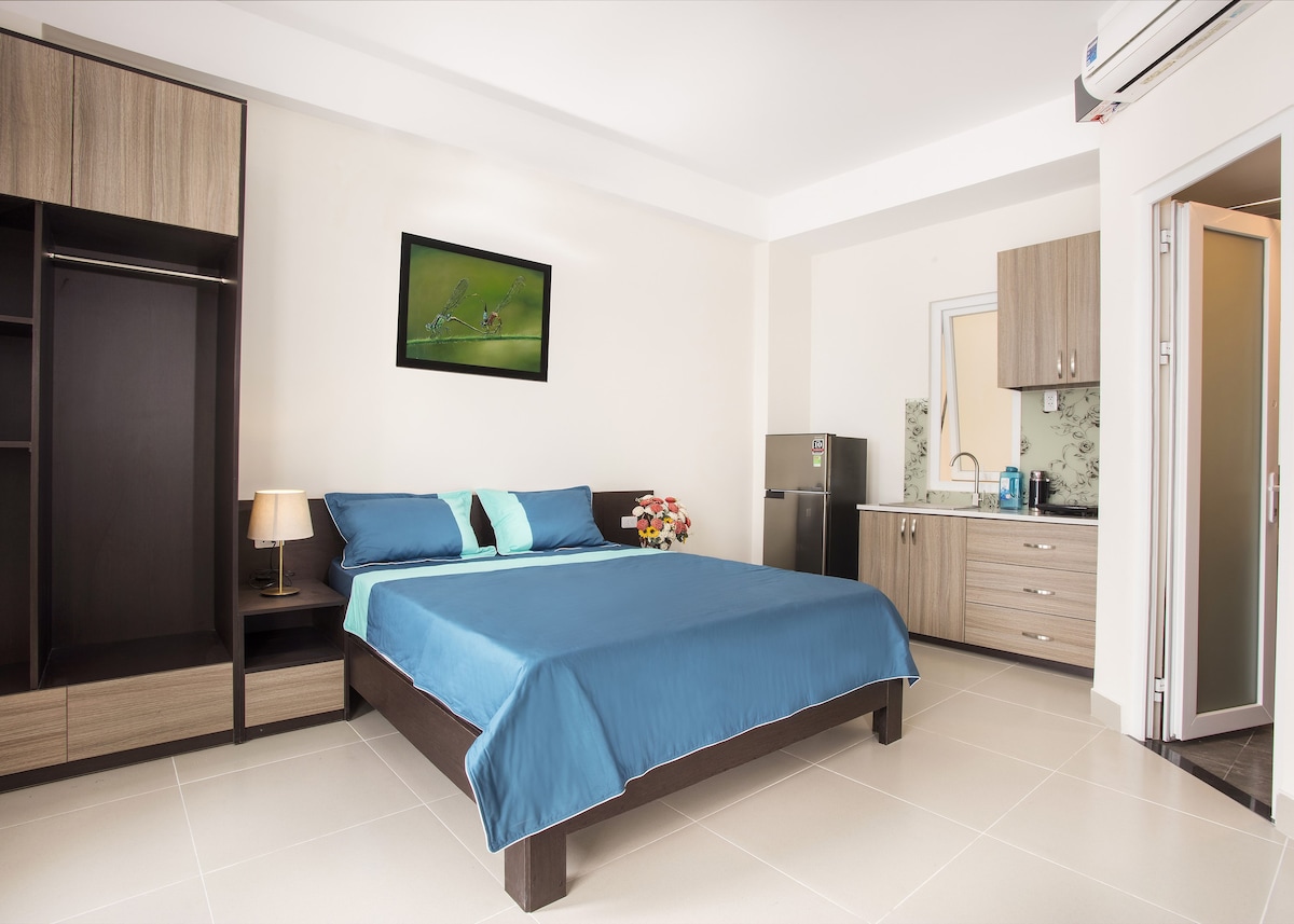 A bright and airy space features a spacious bed with a blue coverlet and contrasting pillows. A kitchenette is visible against the wall, accompanied by a refrigerator. Closets and a bedside lamp provide convenience, while a window allows natural light into the room.