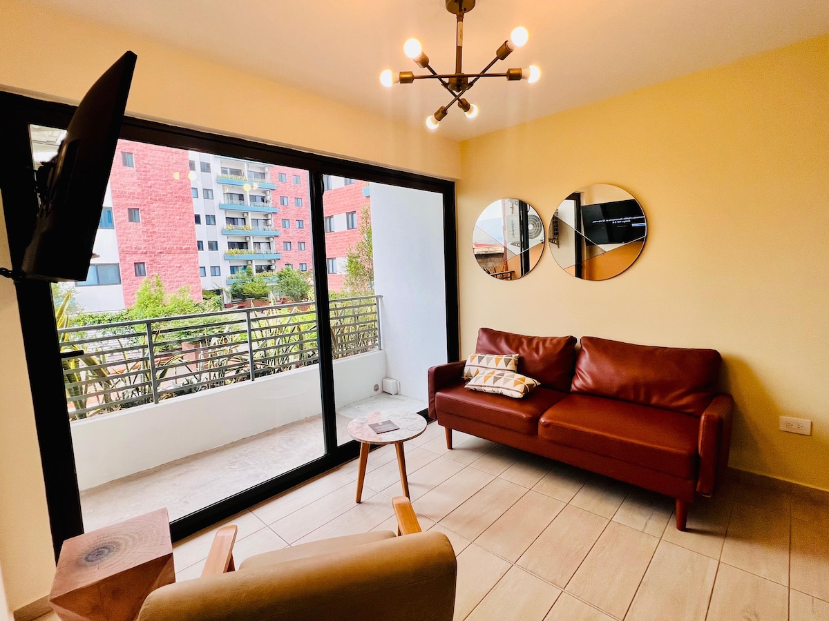 A bright living area features a modern leather sofa positioned near large sliding glass doors. Two circular mirrors adorn the wall, reflecting natural light. A small round table is placed beside the sofa, with a chair visible in the corner, enhancing the inviting setup.