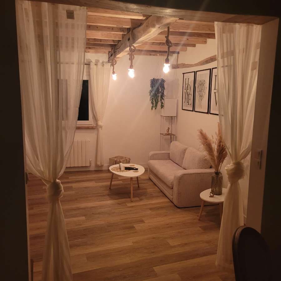 A light-filled living area is presented with wooden beams and soft drapes framing the entrance. A neutral-toned sofa is positioned opposite a small round coffee table topped with decorative items. Artistic prints adorn the walls, and a warm ambiance is created by hanging light fixtures.