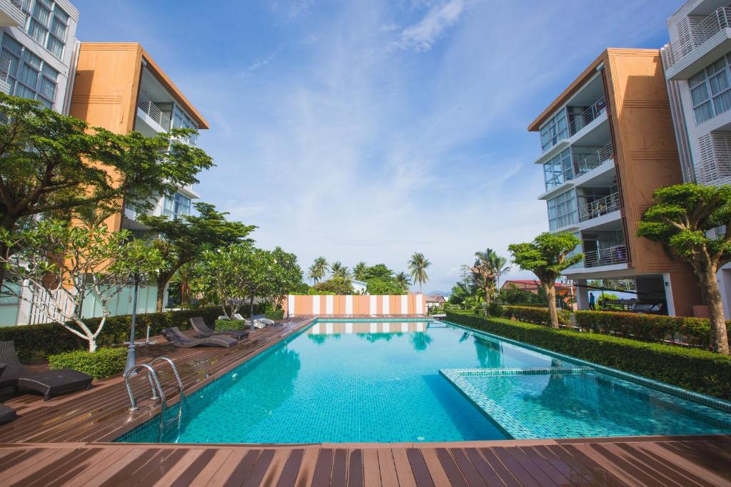 A clear view of a spacious saltwater swimming pool is presented, surrounded by well-maintained greenery and wooden decking. Lounge chairs are positioned along the poolside, with modern apartment buildings visible in the background under a bright blue sky.
