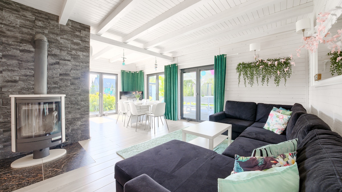 A spacious living area features a large, dark sectional sofa facing a sleek white coffee table. A wood-burning stove is positioned against a textured stone wall. Bright green curtains frame the windows, allowing light to illuminate the room. A dining table is visible in the background.