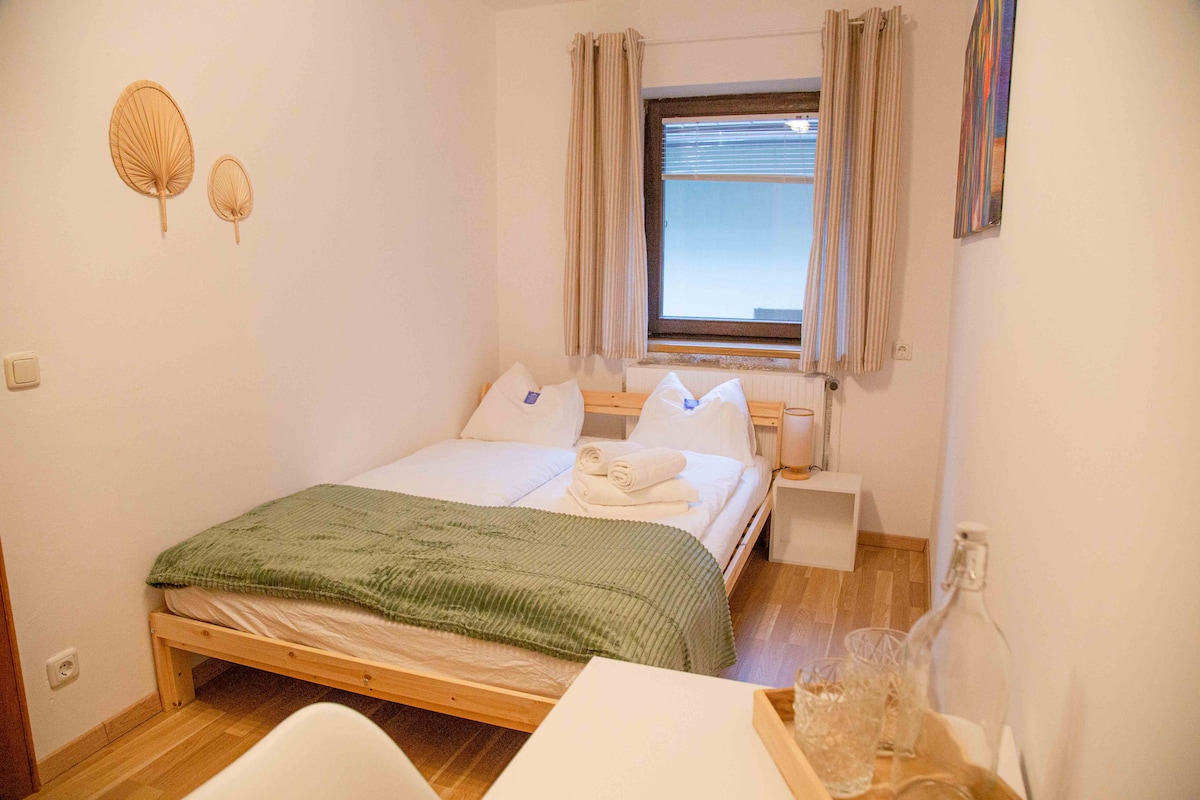 A bright sleeping area features a large bed with two pillows and a green throw. Natural light enters through a large window, complemented by light curtains. Minimalist decor is present, including decorative wall elements and a small table set with glassware.