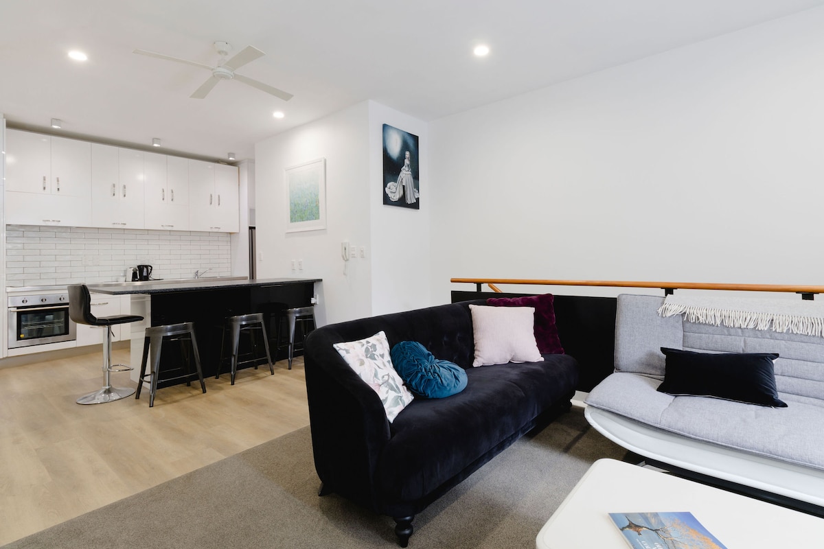 A modern living area features a dark sofa complemented by colorful cushions. A light-colored coffee table is situated in front. In the background, an open plan kitchen is visible, equipped with sleek cabinetry and bar-style seating at the counter.