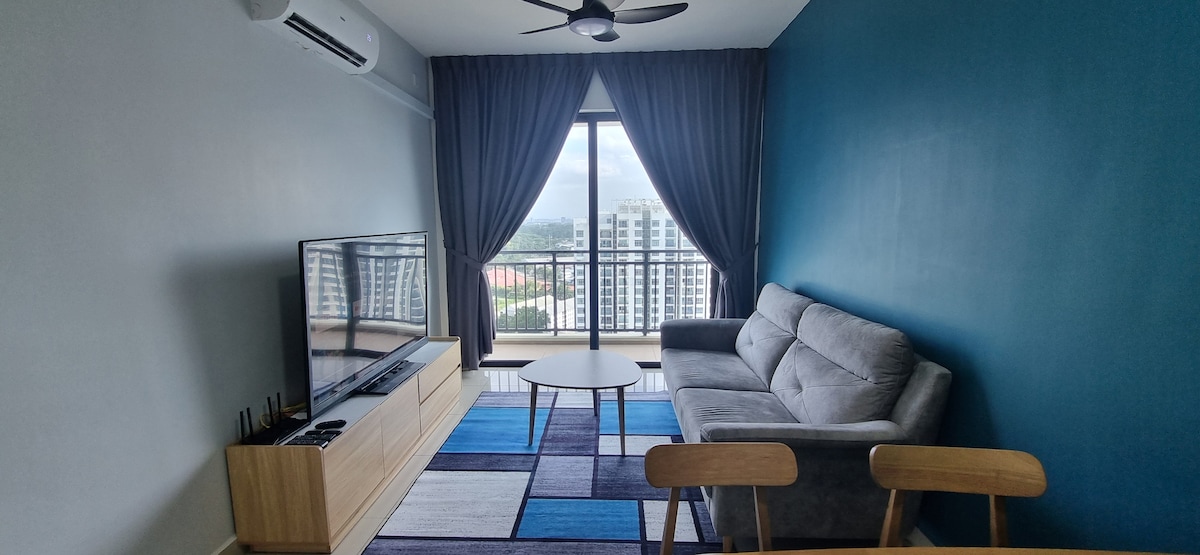 A living area features a gray sofa positioned against a vibrant blue accent wall. Natural light enters through large windows, revealing a city view. A round coffee table sits on a patterned rug, while a flat-screen TV is mounted on a wooden console.