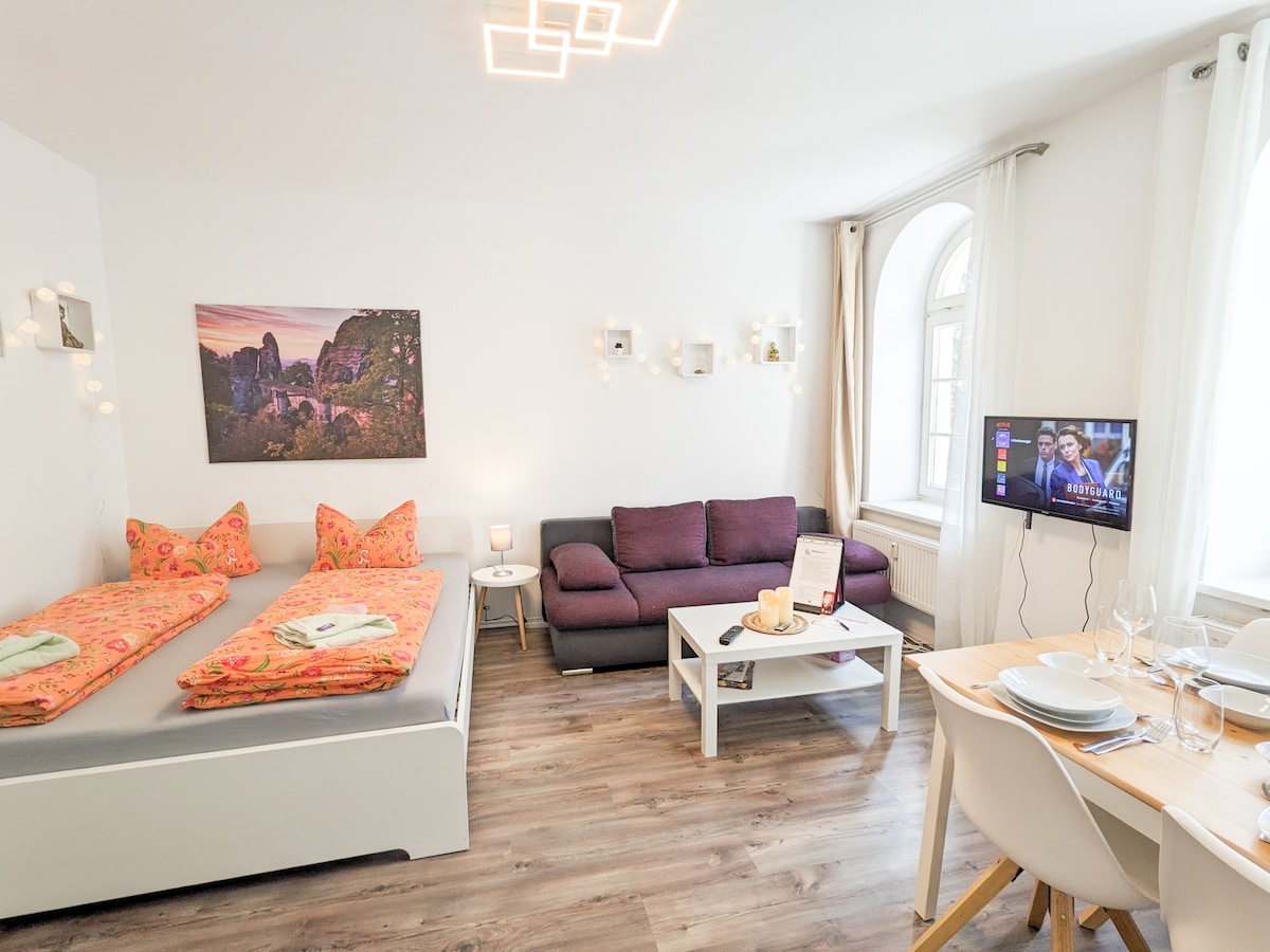 A bright living area features a spacious bed with decorative orange cushions and a cozy sofa in a neutral tone. A small dining table is set with tableware, and a television is mounted on the wall. Light streams in through a large arched window.