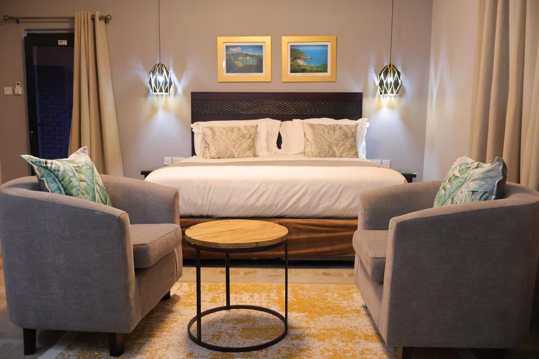 A spacious sleeping area is characterized by a large bed, flanked by modern bedside lamps. Two comfortable armchairs with decorative pillows are positioned near a round wooden table, enhancing the inviting atmosphere. Artwork hangs above the bed, complementing the neutral wall tones.