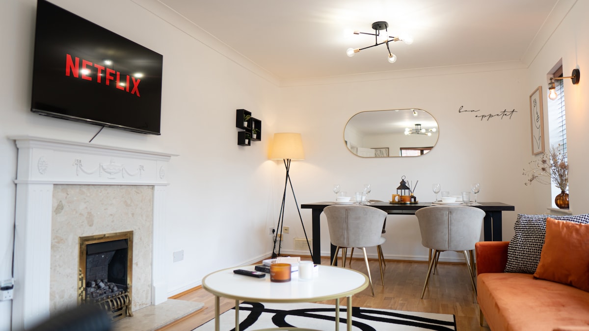 A stylish living area features a modern fireplace and a TV mounted on the wall displaying content. A dining table with gray chairs is set for guests, while a lamp provides gentle lighting. An orange sofa adds a pop of color to the neutral decor.