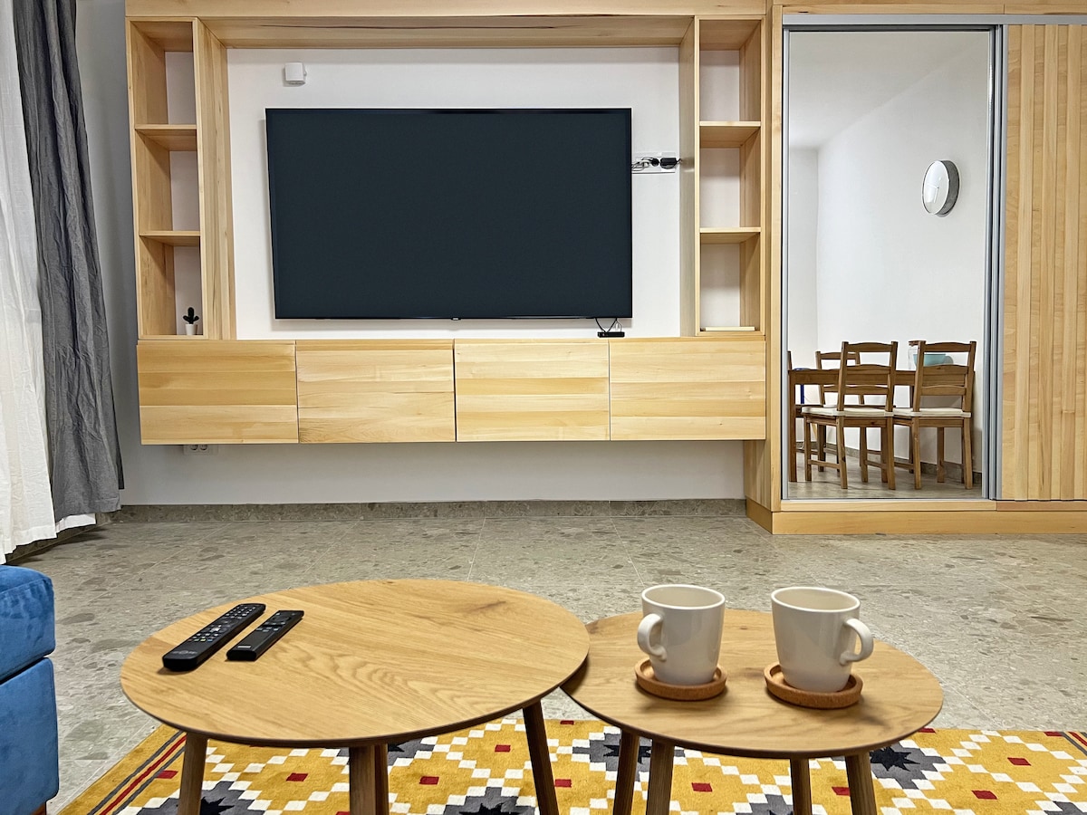 A modern living area showcases a wooden media unit with a large flat-screen TV mounted above it. Two round wooden tables each hold a white cup. The background reveals a dining area with wooden chairs and a mirrored door, enhancing the spacious look.