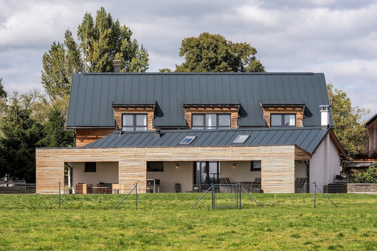 The modern holiday home features a combination of wood and stone exteriors, highlighted by a large covered terrace. Multiple skylights enrich the upper section, while expansive windows provide views of the surrounding grassy landscape. The structure is bordered by a secure fence.