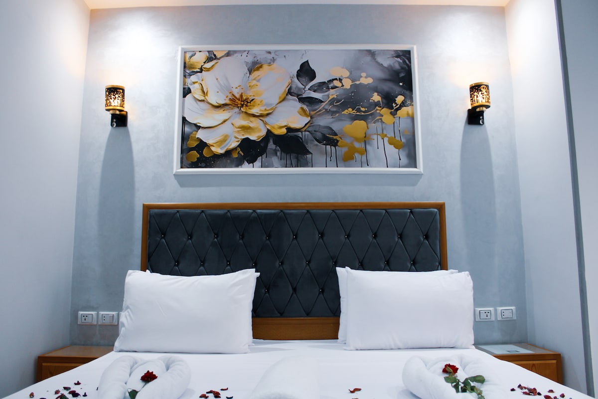 A modern bedroom features a large headboard with a diamond pattern, flanked by two stylish wall sconces. Above the headboard, a floral artwork with gold accents adds a touch of elegance. Freshly arranged towels and scattered rose petals embellish the spacious bed.