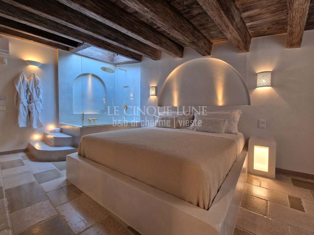 A spacious sleeping area features a large bed with a neutral-toned blanket. Soft lighting is provided by sconces on the curved walls. A cozy seating area is created by steps leading to a raised platform, complemented by bathrobes hanging nearby.