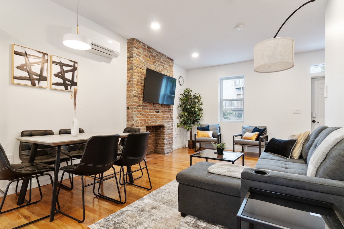 A bright living area features a sectional sofa and a sleek coffee table, surrounded by modern seating. A brick accent wall enhances the space, while a mounted flat-screen TV adds functionality. Natural light enters through a nearby window, creating a welcoming atmosphere.