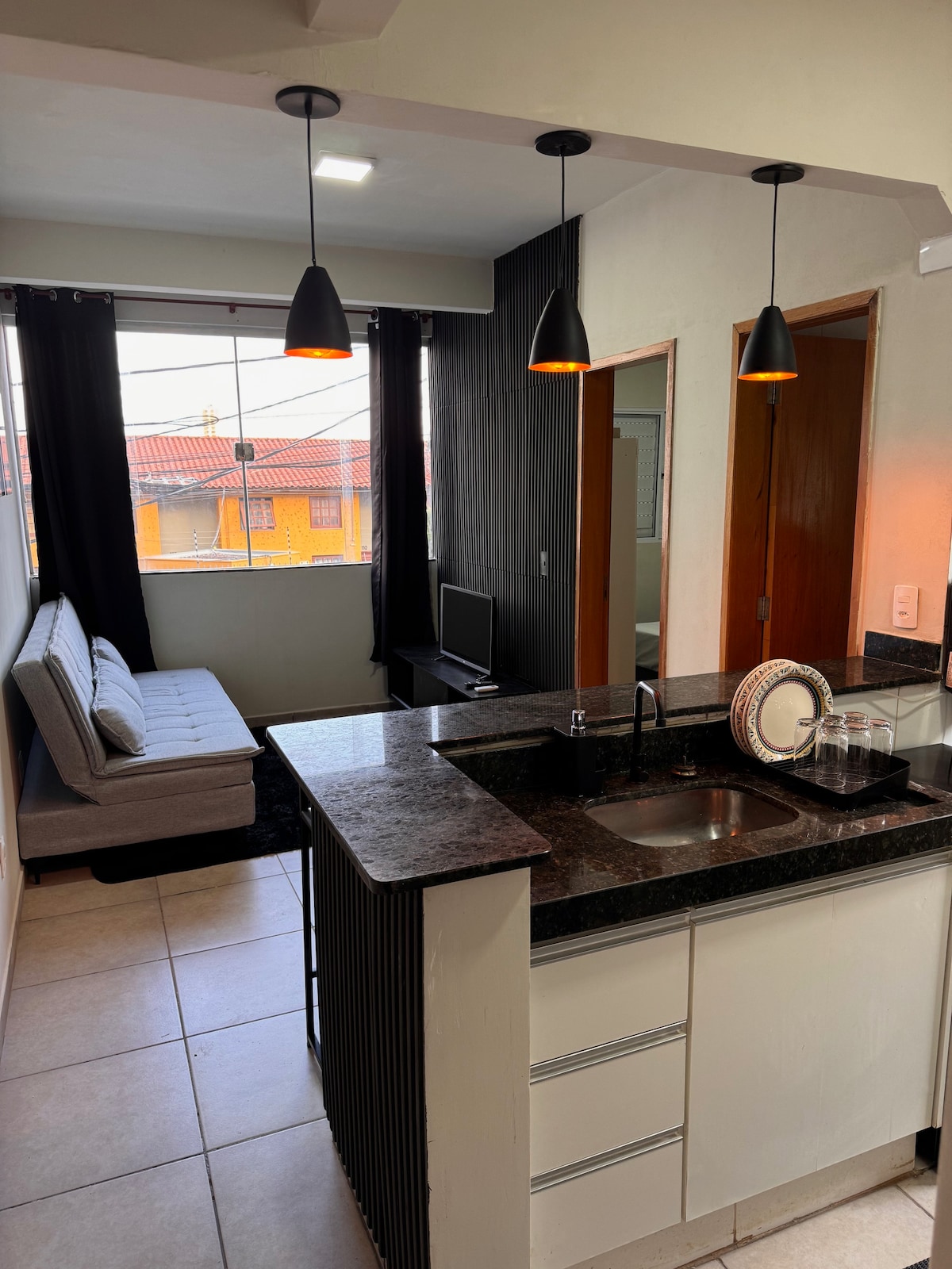 A well-equipped kitchenette with dark countertops is visible, featuring a sink and modern fixtures. Natural light filters through large windows adorned with black curtains. A comfortable sofa bed is positioned beside the kitchenette, and a small television sits on a dark stand against the wall.