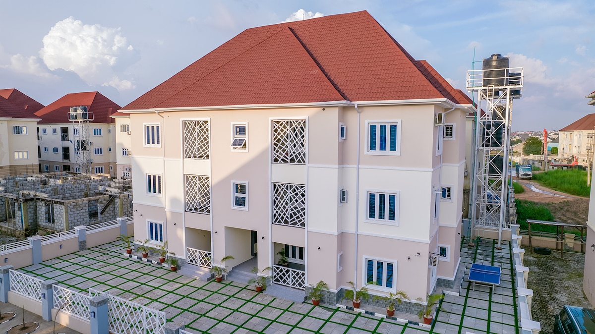 The exterior of a modern three-story apartment building features a light-colored facade with white accents. A neatly arranged courtyard showcases lush greenery and paved pathways, while multiple windows allow natural light to flood the interiors. A shaded area with seating is visible, enhancing outdoor options.
