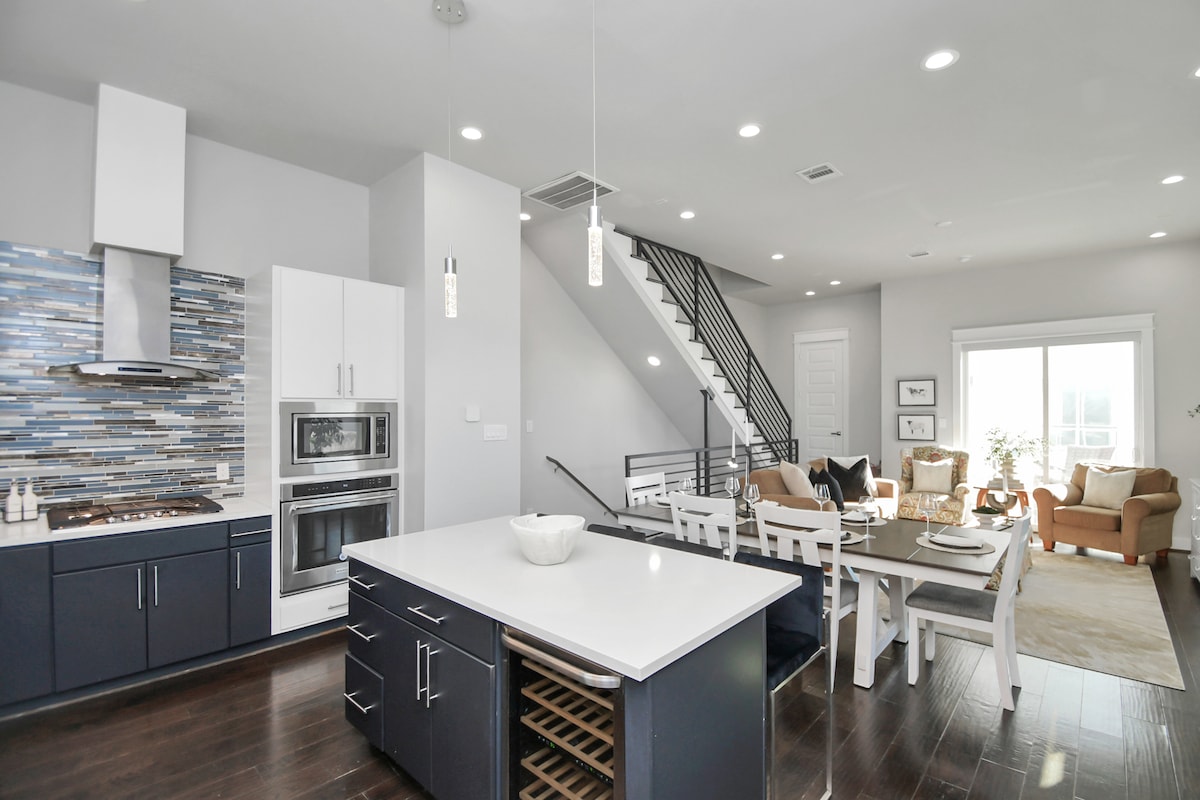 An open concept kitchen and living area features modern appliances, including a stainless steel oven and microwave, set against a stylish backsplash. A spacious island with a wine chiller is accompanied by a dining table and comfortable seating nearby. Natural light floods the room through large windows.