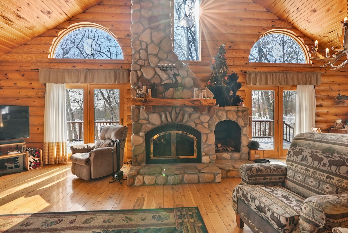 A spacious Gathering Room is highlighted by a floor-to-ceiling river rock fireplace centered against wood-paneled walls. Large windows allow natural light to flood the space, while soft seating arrangements invite relaxation. A plush armchair complements the cozy atmosphere, framed by views of the outdoors.