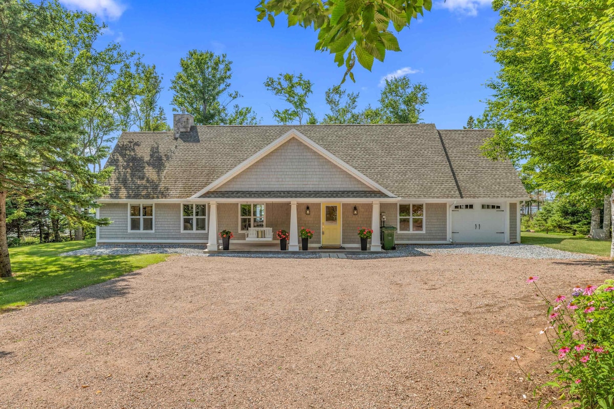 A welcoming exterior features a single-story home with a light gray facade and a charming front porch. Surrounded by lush greenery, the property includes a spacious gravel driveway and flower planters near the entrance, providing a comfortable entry point for guests.