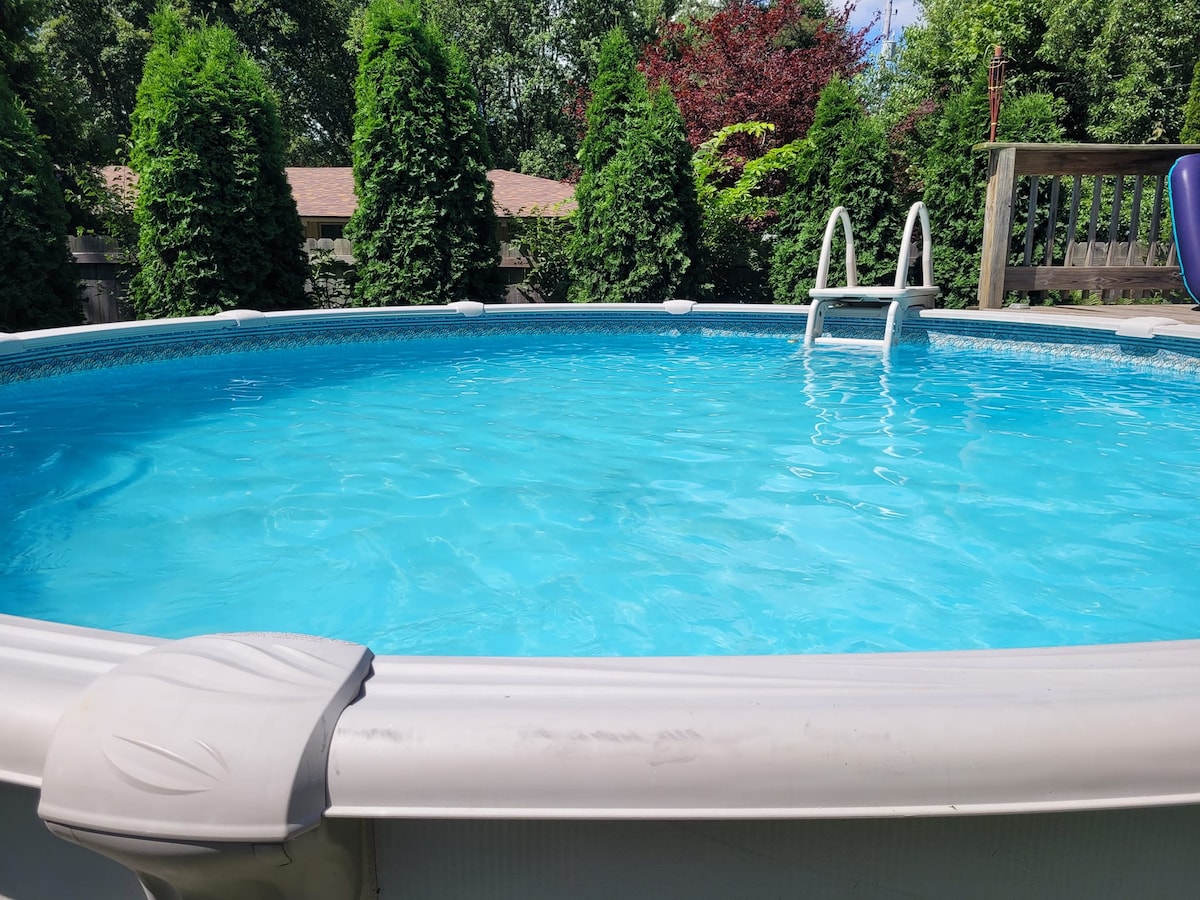 A round swimming pool is filled with clear blue water, surrounded by lush greenery. A white ladder is positioned at the side for easy access. The area reflects natural light and offers a serene outdoor setting.