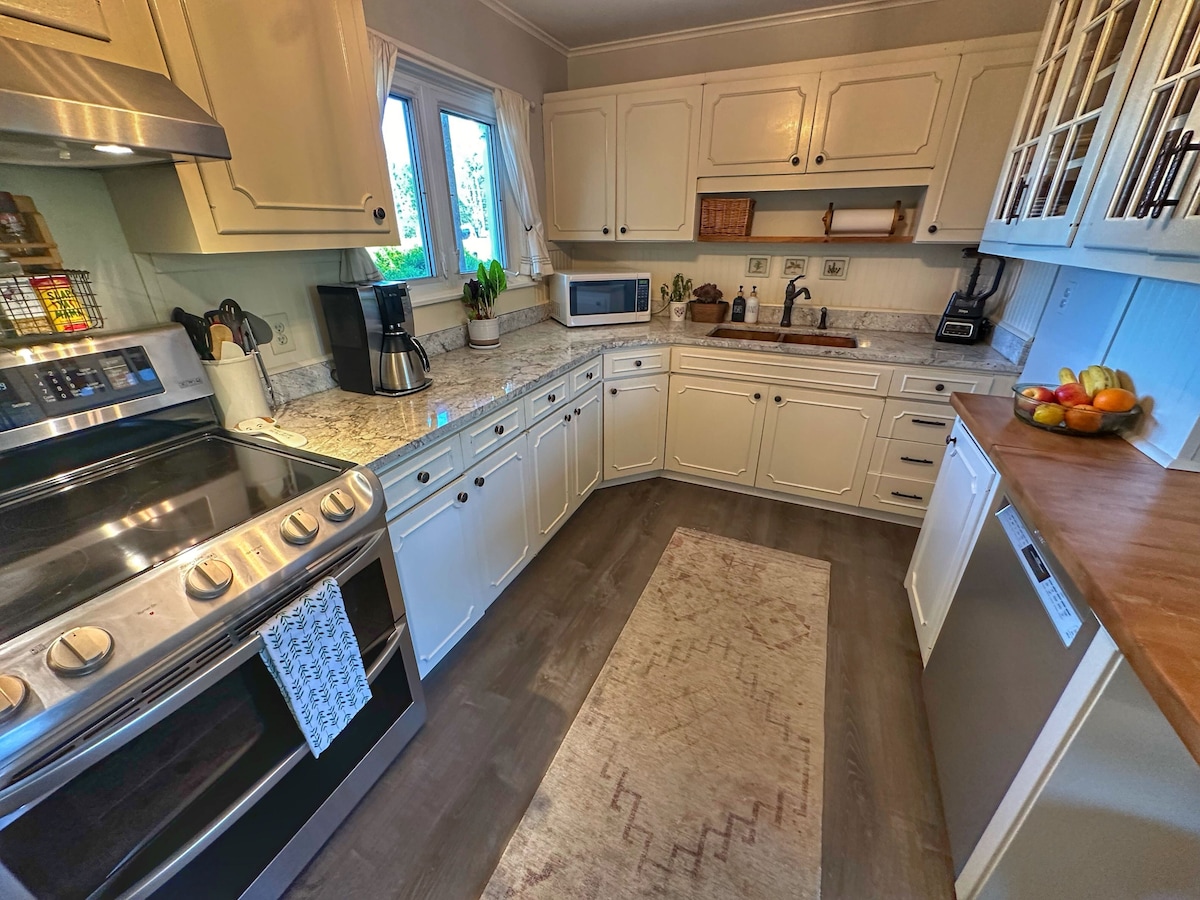 The kitchen features modern appliances and ample counter space, highlighted by a stylish backsplash. A window allows natural light to illuminate the area, complemented by white cabinetry and a wooden countertop. Fresh fruit is displayed in a bowl, adding a touch of color to the setting.