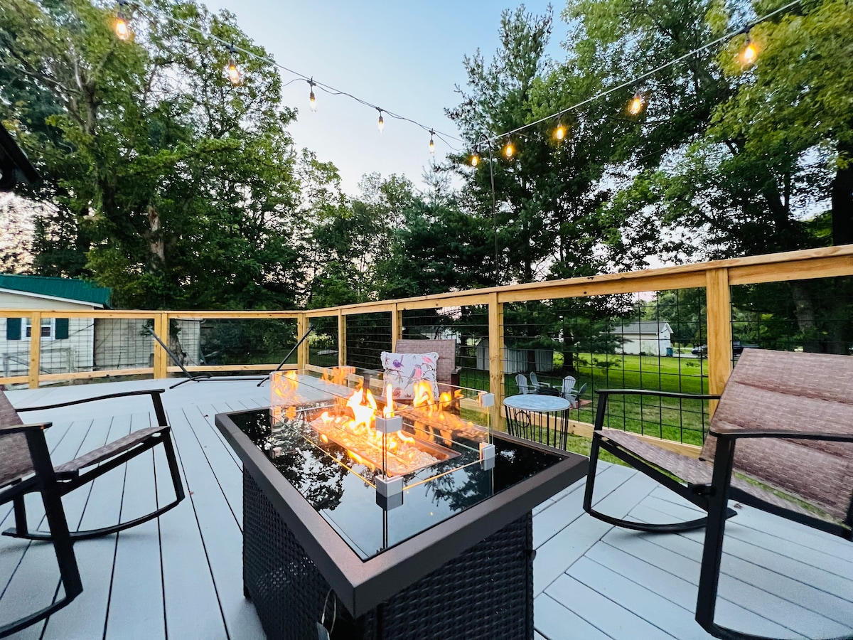 A spacious outdoor deck features a propane fire table surrounded by comfortable seating. String lights hang overhead, enhancing the ambiance during twilight. Lush trees and greenery frame the area, providing a serene backdrop for gatherings or relaxation.