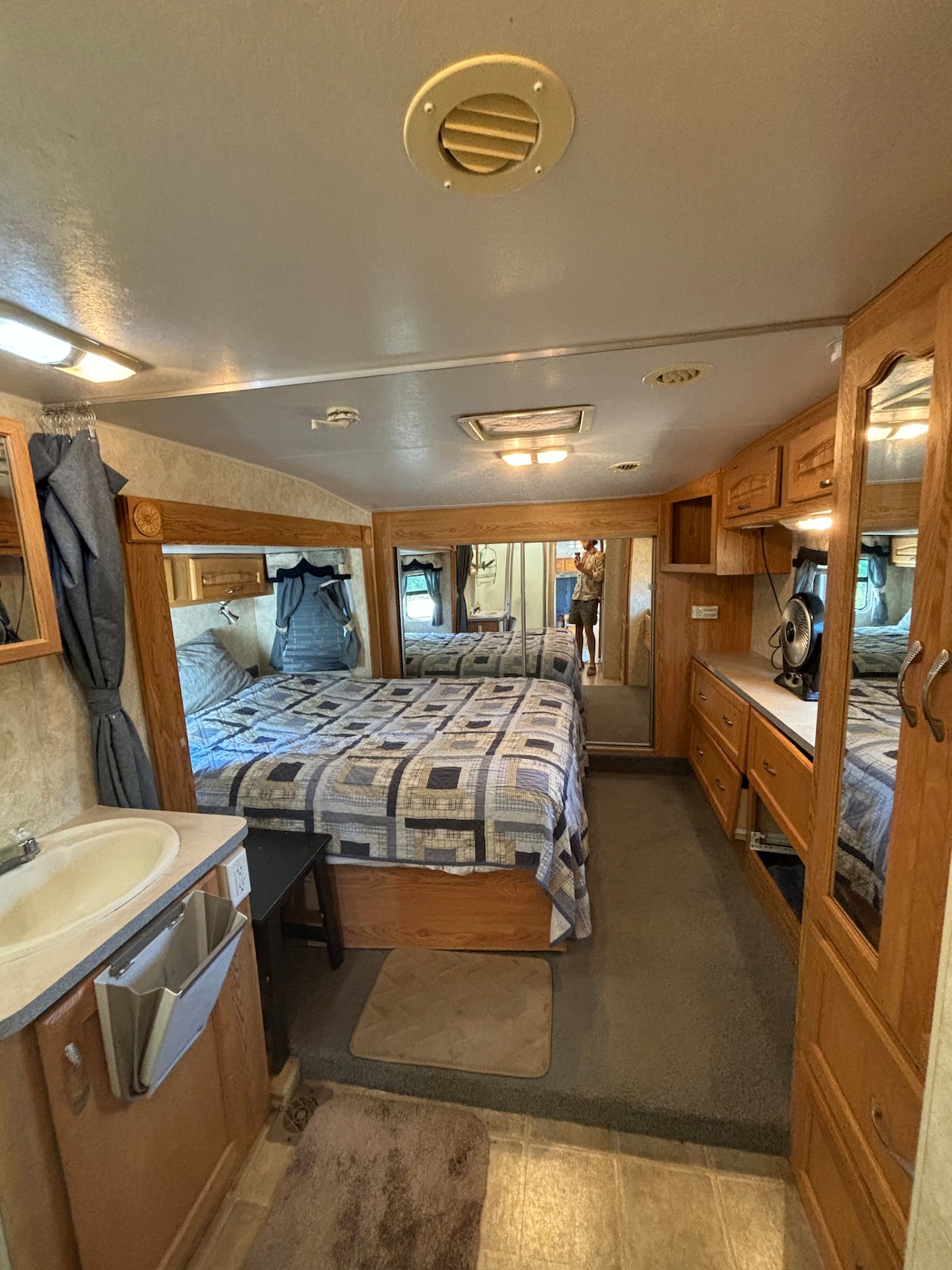 The interior of the camper features a cozy sleeping area with a patterned bedspread and built-in cabinetry. A small sink area is visible beside a mirror with ample natural light. A carpeted floor adds comfort, and the space is well-lit with overhead fixtures.