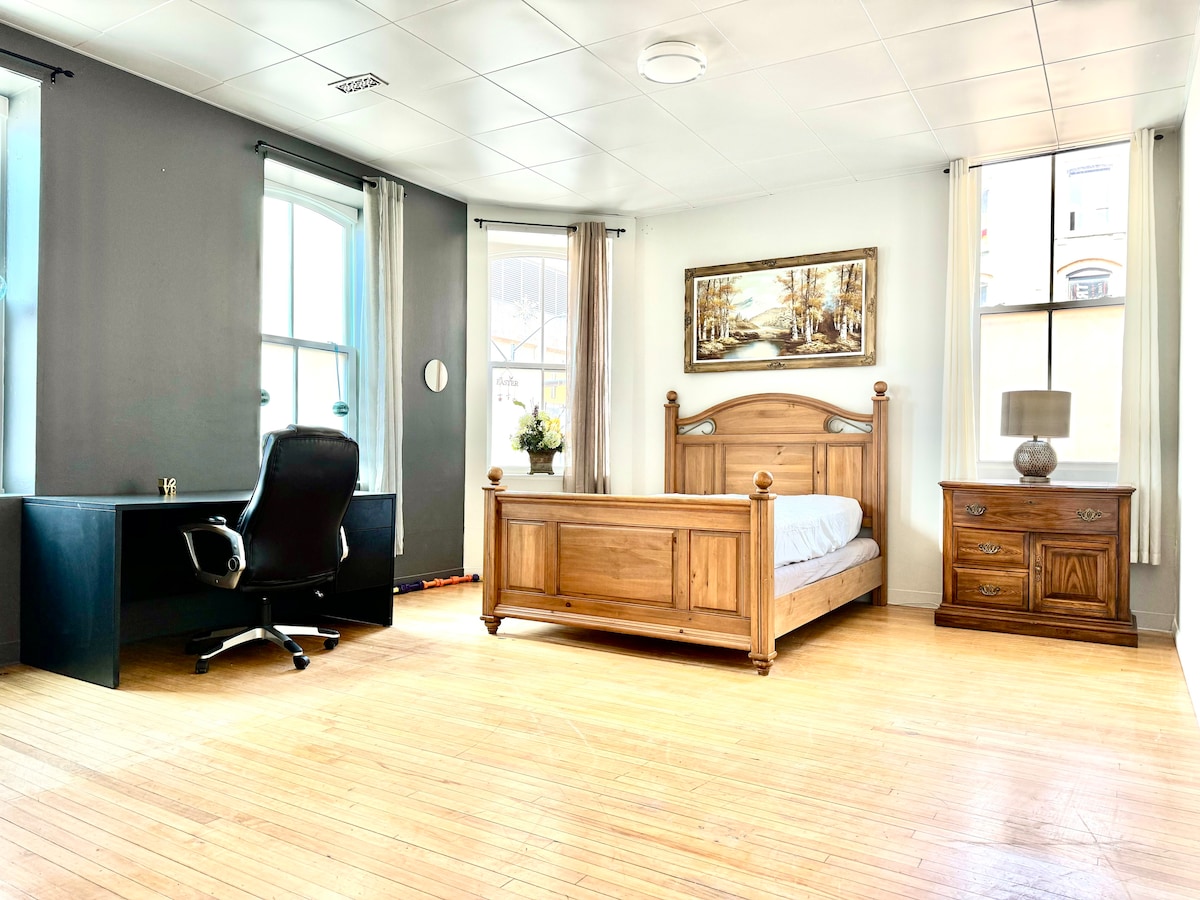 A bright bedroom features large windows allowing ample natural light. The space includes a wooden bed frame and a nightstand, complemented by a desk and an office chair. Simple artwork hangs on the wall, while light-colored curtains frame the windows.