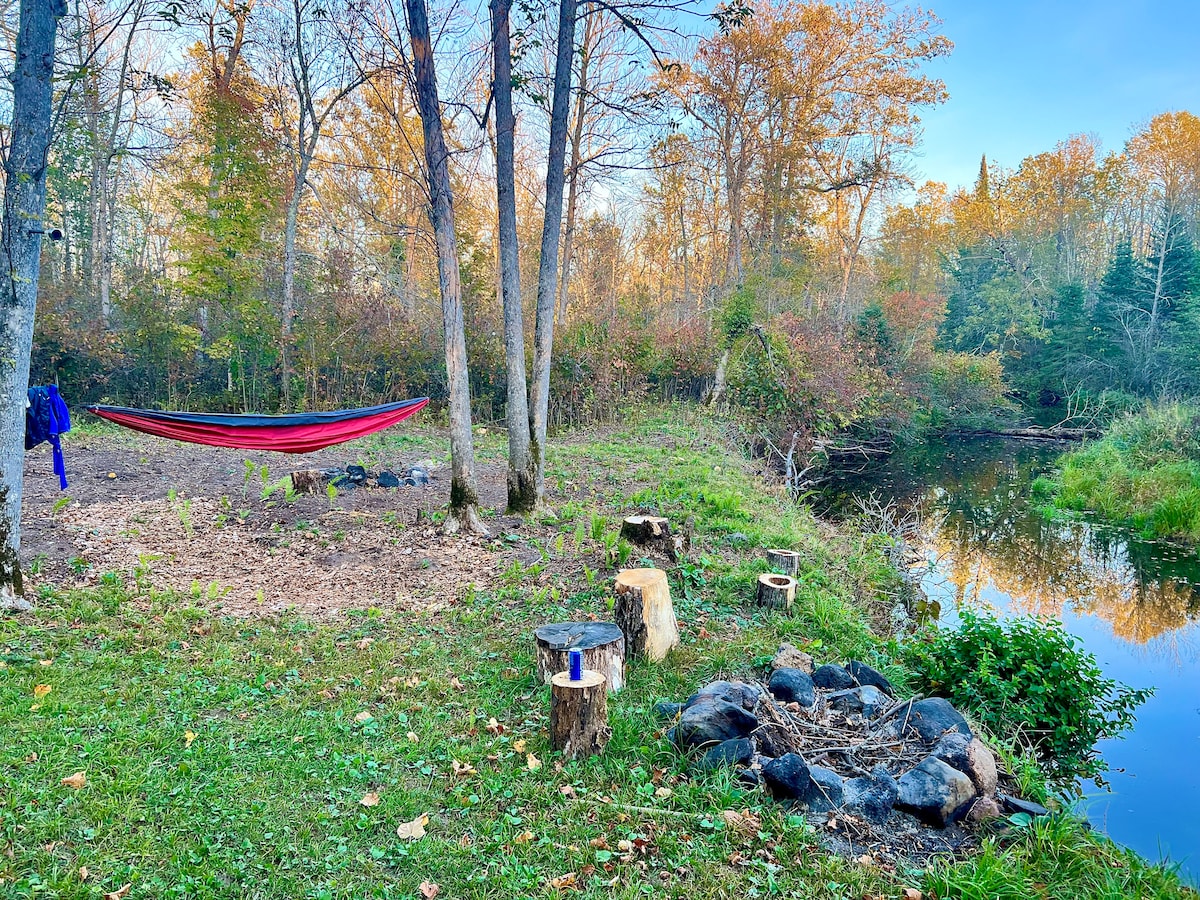 A serene riverside area features a colorful hammock suspended between two trees. The surrounding landscape includes a grassy patch with cut tree stumps and a fire pit composed of dark stones. Autumn foliage adds warm hues to the backdrop along the riverbank.