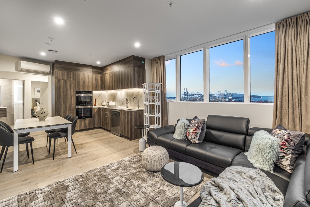 The modern living area features a sleek black couch and a soft area rug, complemented by a round coffee table. A dining table for four sits adjacent to the kitchen, which includes high-end appliances. Large windows provide a view of the waterfront and let in natural light.