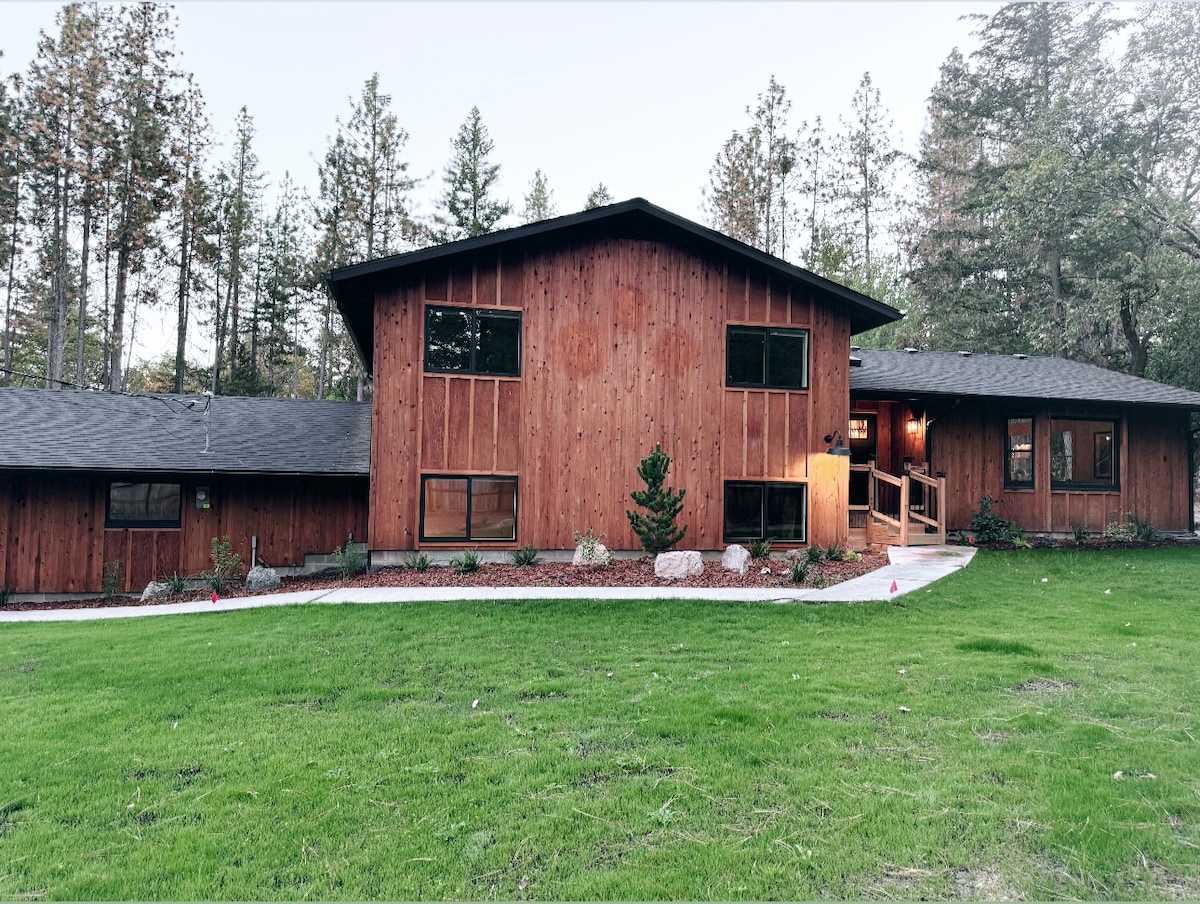 The newly remodeled home features a warm wooden exterior with large windows and a welcoming entrance. A neatly manicured lawn surrounds the house, with a pathway leading to the entrance. The structure is set against a backdrop of tall trees, enhancing the natural setting.