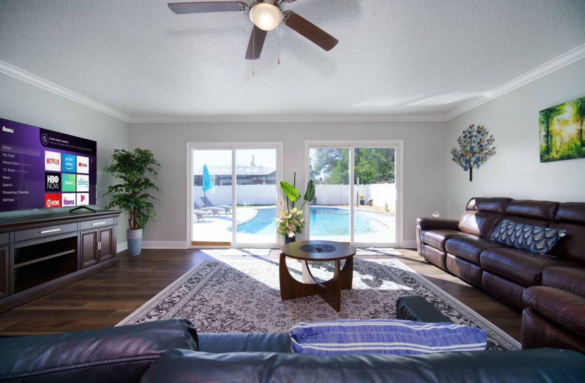 A spacious living room showcases a large TV mounted on a dark entertainment unit. Natural light fills the area through expansive glass doors, which provide a view of the private pool. Comfortable leather sofas surround a circular coffee table, enhancing the relaxed ambiance.