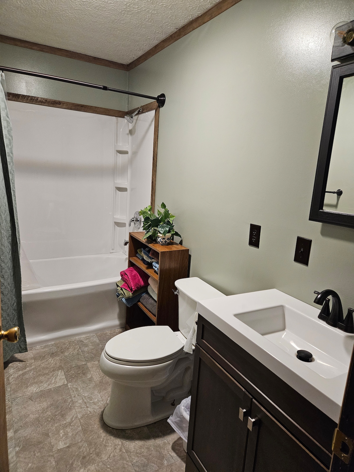 A well-appointed bathroom features a clean, white bathtub with a shower curtain. A dark wooden shelf holds neatly folded towels and decorative items. A modern vanity with a sleek sink and mirror is complemented by subtle green walls and soft lighting.