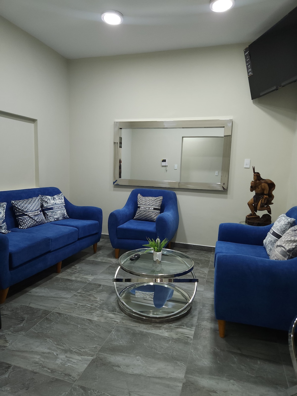 A seating area is showcased with two blue sofas and a matching armchair, all adorned with decorative pillows. A round glass coffee table sits at the center, complemented by a small potted plant. A mirror reflects the space, while a wall-mounted television adds functionality.
