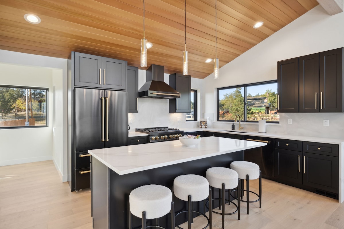 The kitchen features a modern design with black cabinetry and white countertops. An island with seating for four is positioned centrally, under pendant lighting. Large windows provide natural light and showcase views of the outdoors. Stainless steel appliances are integrated seamlessly into the layout.