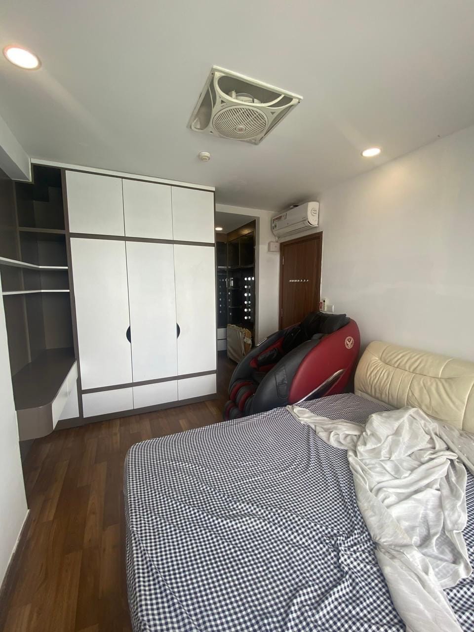 A cozy bedroom features a spacious bed with checkered bedding. An air conditioning unit is mounted on the wall, while a wardrobe provides ample storage. A massage chair sits nearby, and wooden flooring enhances the room's warm ambiance.